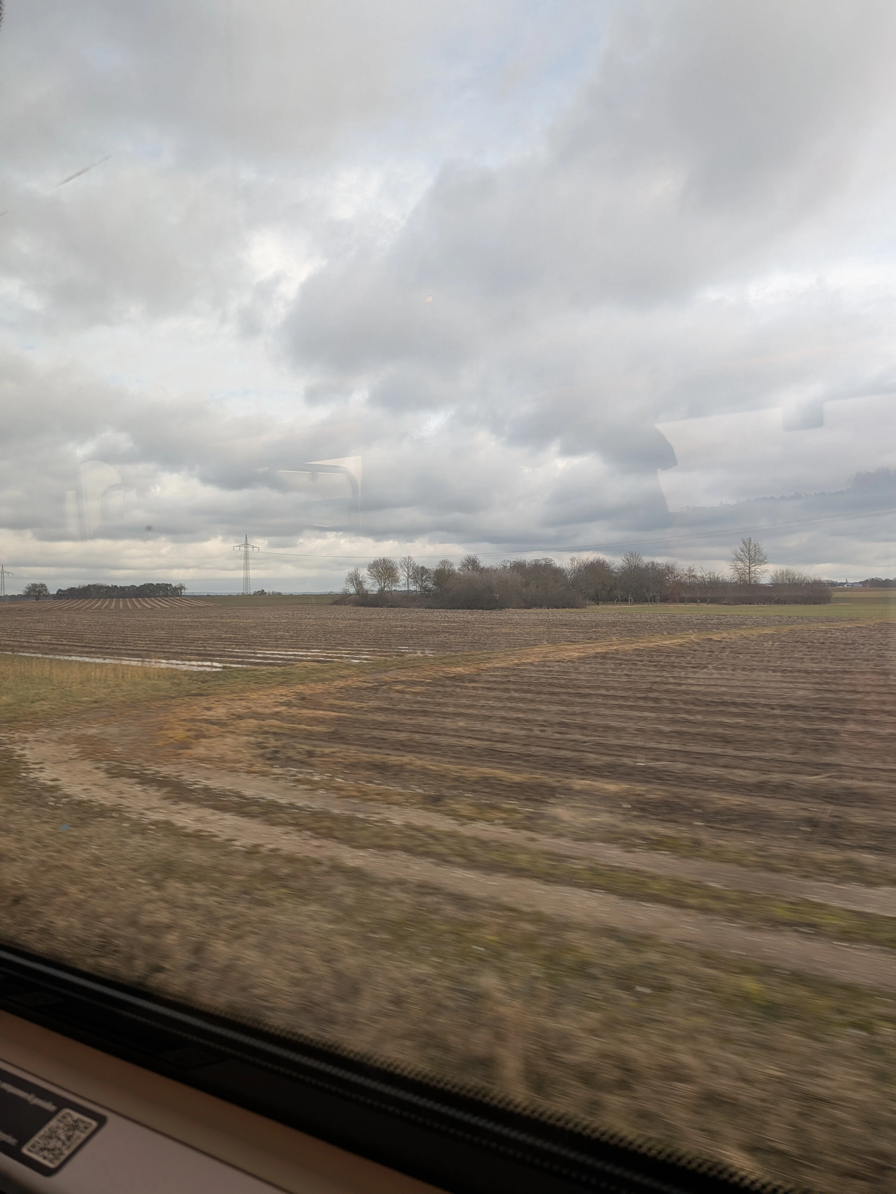 Aussicht aus dem fahrenden Zug. Man sieht eine Landschaft, hauptsächlich Acker mit etwas Gebüsch.